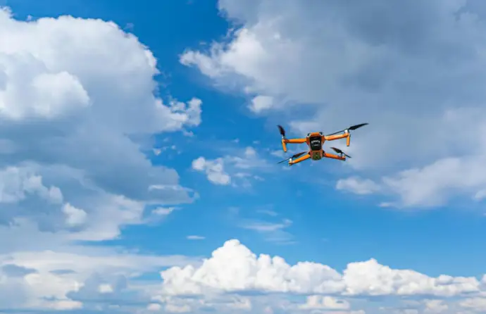 Imagem de um drone amarelo a voar no céu azul com algumas núvens